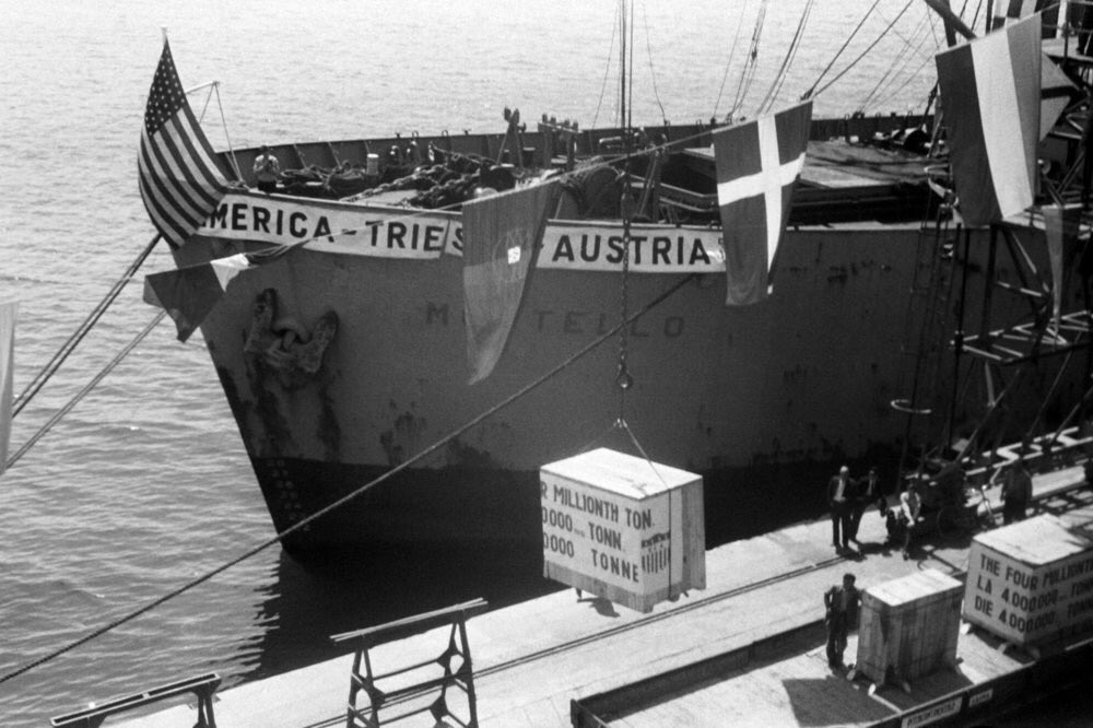 Photo of the four millionth ton. of US aid arriving in Europe. The ship that transported it has a banner reading "America - Trieste - Austria" on the side, and the flags of many Marshall Plan States fly on the foreground. The international Free Port of Trieste played a key role in the reconstruction of Central Europe.